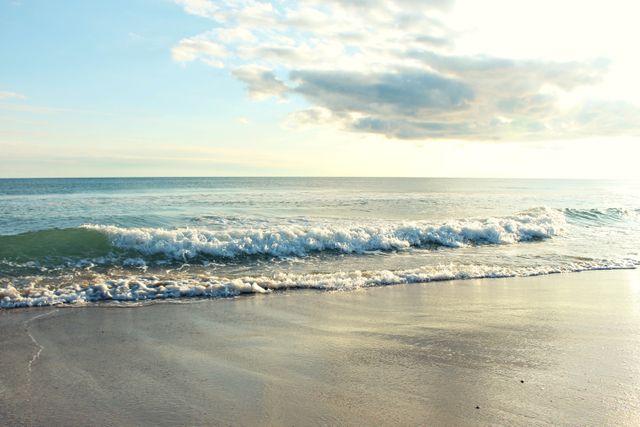 The image shows gentle ocean waves washing ashore with a clear horizon under a setting sun. Minimal clouds are present in a bright sky, creating a peaceful atmosphere. Ideal for travel blogs, relaxation or meditation content, and beach-themed promotions.