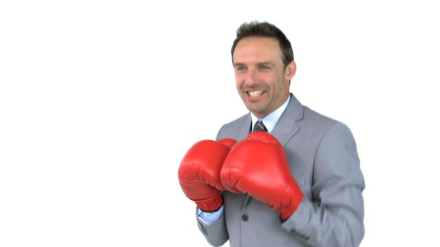 Businessman in a grey suit and red boxing gloves smiling and throwing punches in front of a white background. Suitable for themes related to office fitness, corporate wellness, determination, and blending professional work with physical activity. Can be used in promotional materials, blog posts, and advertisements showcasing positive work-life balance, stress-relief techniques, or motivation in business settings.