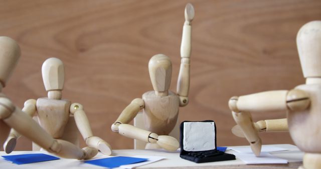 Wooden art mannequins sitting around a table with a laptop, appearing engaged in learning and discussing. One figure is raising its hand as if asking a question, while others are positioned in attentive or collaborative stances. Useful for representing concepts of teamwork, brainstorming sessions, creativity, education, and collaborative work in an artistic or simplified manner.