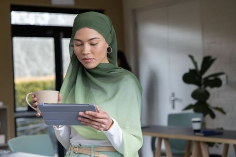 Asian professional in green hijab using tablet and holding mug in modern office lounge