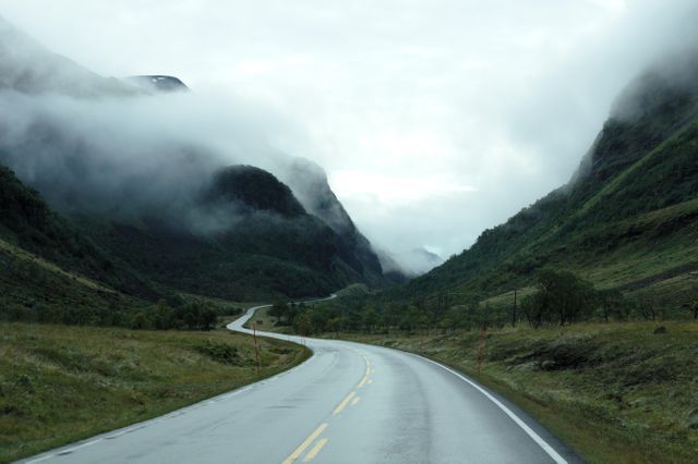 Winding road disappearing into foggy mountain pass, creating ethereal and serene atmosphere. Perfect for travel blogs, adventure campaigns, nature exploration, or backgrounds in presentations and promotions focusing on scenic routes or serenity.