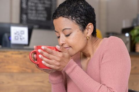African american woman enjoying coffee in cozy cafe holding red mug, smiling and relaxed