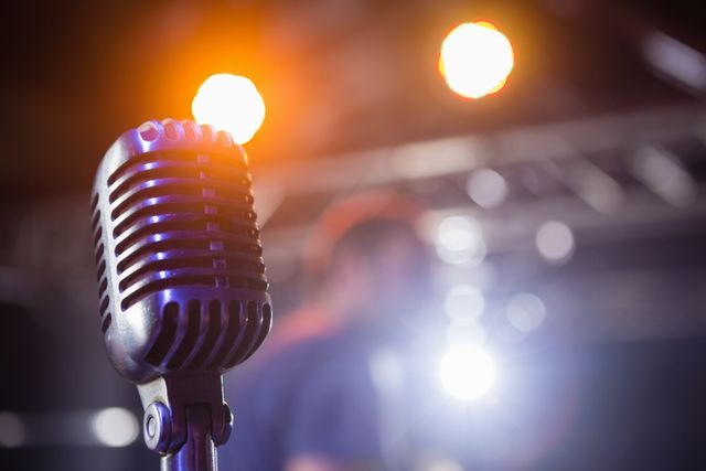 Close-up of a retro microphone on stage with bright lights in the background. Ideal for use in music-related content, concert promotions, event advertisements, and articles about live performances or vintage audio equipment.
