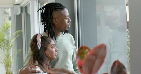 Interracial couple gazing through sunlit window with indoor plants, cozy home moment