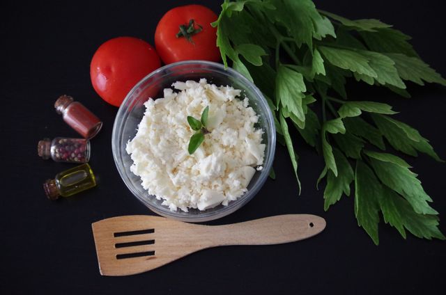 Close-up of fresh feta cheese in clear container garnished with small leaves, surrounded by vibrant tomatoes, fresh herbs, and small bottles of spices on dark background. Perfect for cooking blogs, recipe publications, and healthy eating promotions highlighting fresh ingredients.