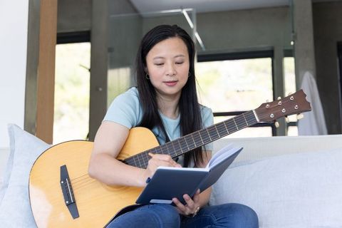 African american woman composing music on acoustic guitar while writing notebook at home