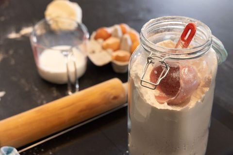 Glass jar of flour with red scoop and rolling pin on dark countertop for baking