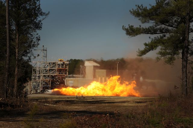 A vibrant scene of a rocket engine test being conducted at an outdoor facility during daytime, showcasing a large fireball and the industrial structures involved. This dynamic image can be used for educational materials on space exploration, engineering advances, demonstrating technological power and breakthroughs in aerospace engineering.