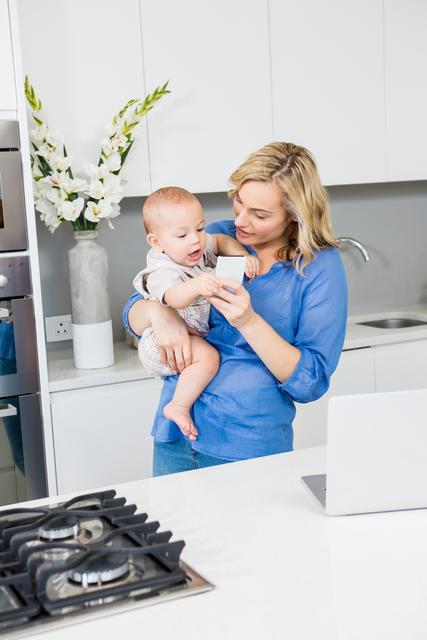 Mother holding baby boy while using mobile phone in modern kitchen. Ideal for parenting blogs, family lifestyle articles, technology in daily life, and home decor inspiration.