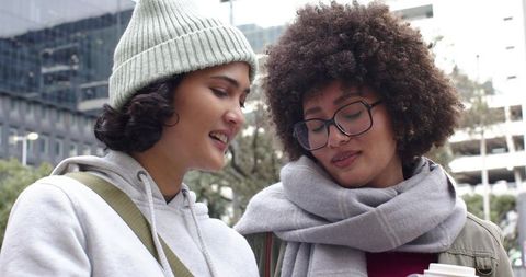Two Friends Sharing Coffee and Laughing on Urban Sidewalk with Winter Accessories