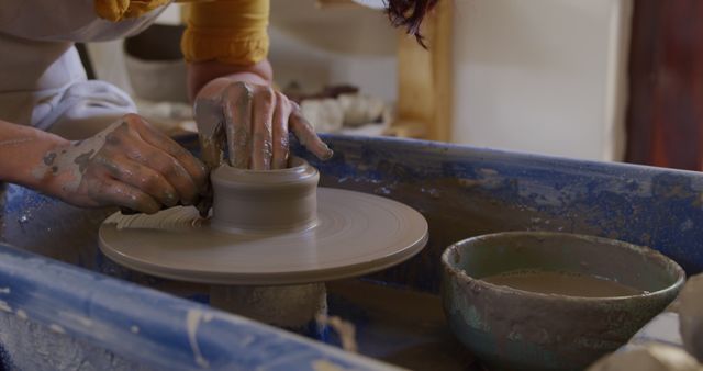 Potter shaping clay on a pottery wheel in a workshop, highlighting the artistic process and craftsmanship. The artisan’s hands are covered in clay, showing the intricate detail work involved in pottery making. This stock photo can be used for creative industry publications, artisan craft advertisements, or educational materials about traditional ceramic techniques.