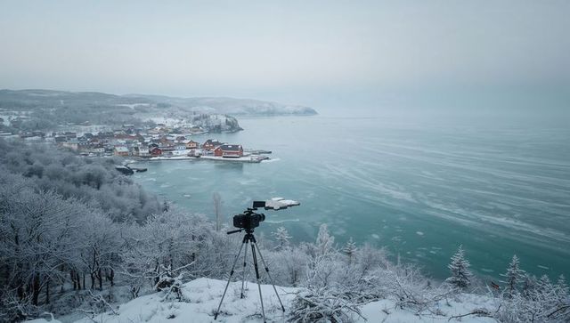 Capturing snowy coastal village from cliff with camera on tripod overlooking frozen bay