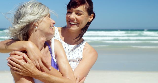 A senior woman and a young adult woman are embracing joyfully on a sunny beach, with copy space. Their laughter and affectionate hug convey a warm, intergenerational family bond.
