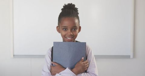 Smiling schoolgirl holding binder by whiteboard