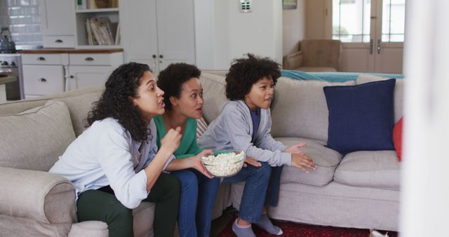 This image shows a family sitting on a couch in a cozy living room, engaging in watching TV while eating popcorn. Ideal for use in advertisements for TV shows, family time, home entertainment systems, or articles promoting spending quality family time at home.
