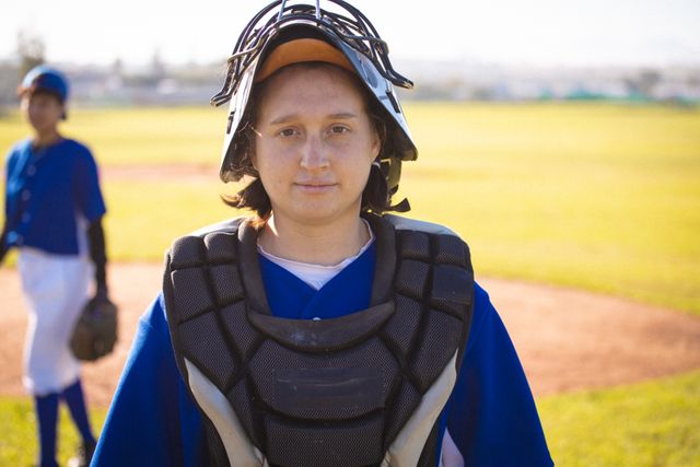 Caucasian female baseball player wearing catcher's gear and helmet, standing on a baseball field with a teammate in the background. Ideal for use in sports-related content, youth sports promotions, team spirit campaigns, and athletic training materials.