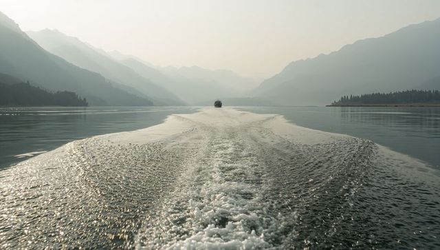 Boat cutting foamy wake across misty mountain lake with layered ridges and forested shore