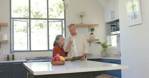 Senior asian couple using tablet in bright modern kitchen with marble island and plants