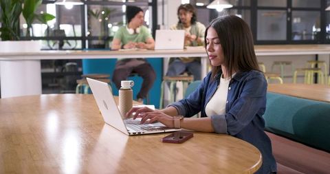 Young woman typing on laptop in modern coworking space, colleagues collaborating behind