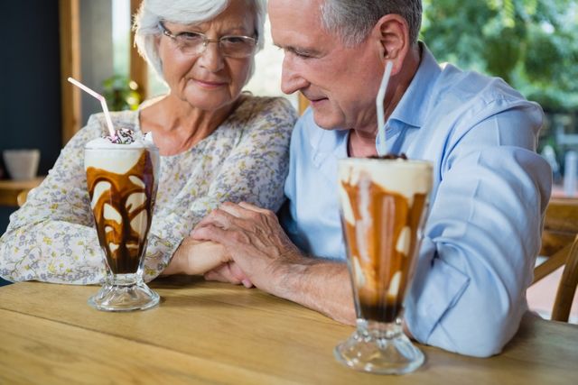 Senior couple holding hands while enjoying coffee and milkshakes in a cozy cafe. Perfect for illustrating themes of love, companionship, and the joys of retirement. Ideal for use in advertisements, blogs, and articles focused on senior living, relationships, and lifestyle.