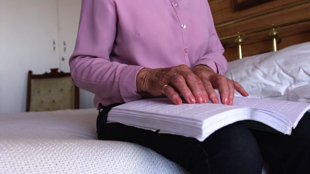 Visually impaired elderly woman wearing a pink blouse is reading a braille book while sitting on a bed in a cozy bedroom. This video can be used for topics on accessibility, senior care, disability awareness, independence in the elderly, inclusion, and educational purposes.