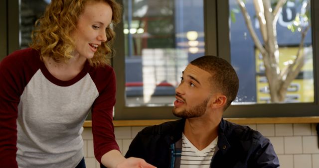 Two friends, a woman with curly hair and a man with a beard, are seen engaging in a lively conversation inside a modern coffee shop. The casual setting suggests a relaxed atmosphere, perfect for socializing and catching up with friends. This image can be used for promoting social interaction, friendship, modern lifestyle, and diverse communities in advertisements, blogs, and social media content.