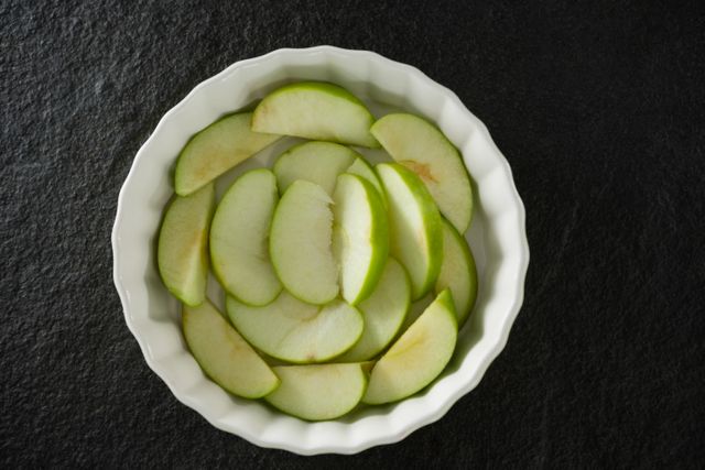 This image shows green apple slices neatly arranged in a white bowl from an overhead perspective. It is ideal for use in food blogs, recipe websites, healthy eating promotions, and kitchen-related content. The fresh and vibrant appearance of the apple slices makes it suitable for illustrating concepts of nutrition, organic food, and vegan or vegetarian diets.