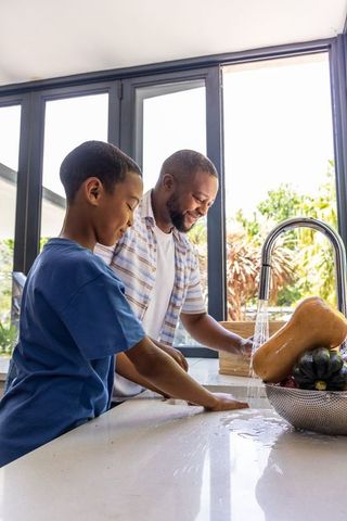 African american father and teenage son rinsing vegetables at sunlit modern kitchen island