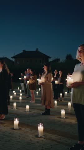 Vertical video showing community standing holding candles in organized vigil grid at night