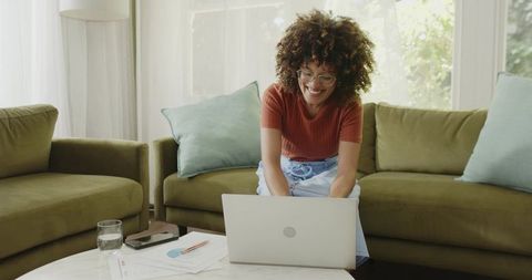 African American woman working from home on laptop reviewing papers at sofa coffee table