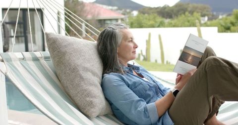 Mature woman reading on striped hammock by pool, relaxing with smartwatch and sunset paperback