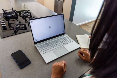 Mature woman leaning at kitchen counter using laptop and notebook for remote work