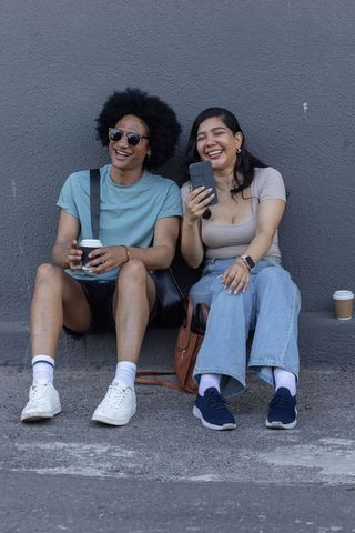 Laughing diverse friends sharing smartphone and coffee sitting on urban concrete ledge