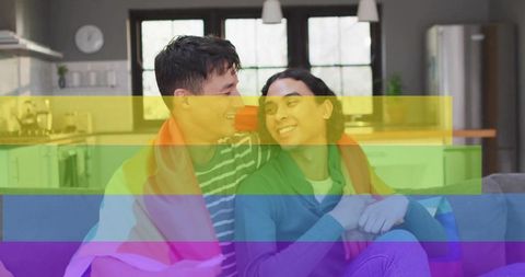 Couple cuddling under rainbow pride flag on modern sofa, smiling and embracing at home