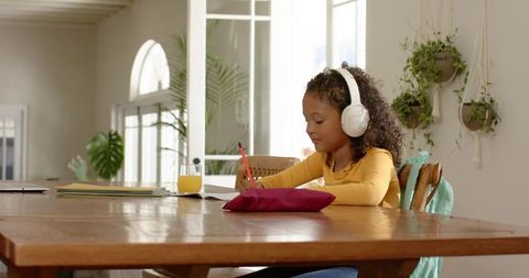 African american girl studying at table with headphones writing homework in sunlit room