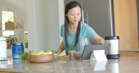 Asian woman preparing healthy smoothie at kitchen island while following recipe on tablet