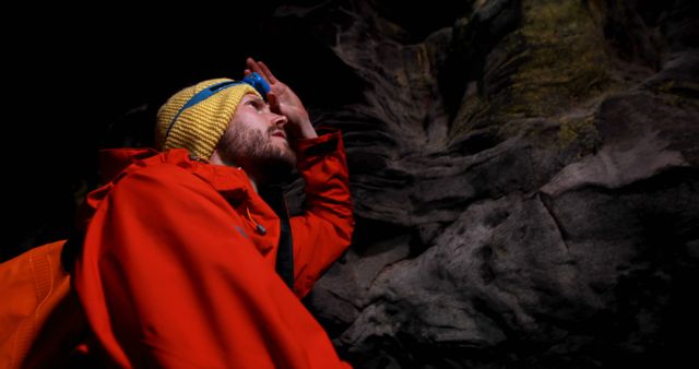 Male hiker in a bright orange jacket and yellow beanie holds flashlight searching in dark cave. Useful for articles on outdoor activities, adventure tourism, safety gear, and camping equipment. Ideal for inspirational travel blogs, extreme sports promotions, and nature exploration guides.