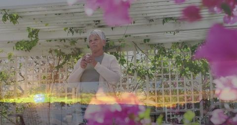 Senior woman standing on pergola-covered terrace holding cup among pink blooms at sunset