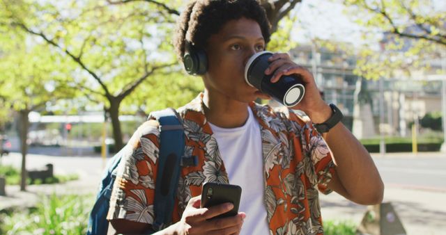 Young man enjoying a casual walk, engaging with his smartphone while drinking coffee and wearing headphones offers great content for lifestyle blogs, social media promotions for cafes, and urban living adverts.