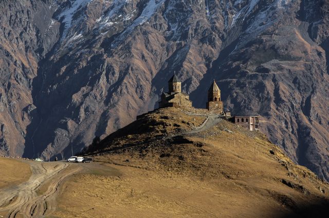 Gergeti Trinity Church situated high on a mountain ridge with a dramatic rocky mountain backdrop. The historic architecture contrasts beautifully with rugged natural scenery. Perfect for uses in travel guides, cultural articles, tourism advertisements, and religious studies material.