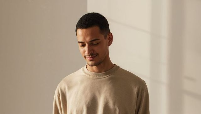 Serene young man standing in beige sweatshirt gazing downward with warm window shadows