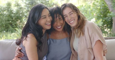 Multiracial women laughing and hugging on sunlit outdoor sofa, candid friendship moment