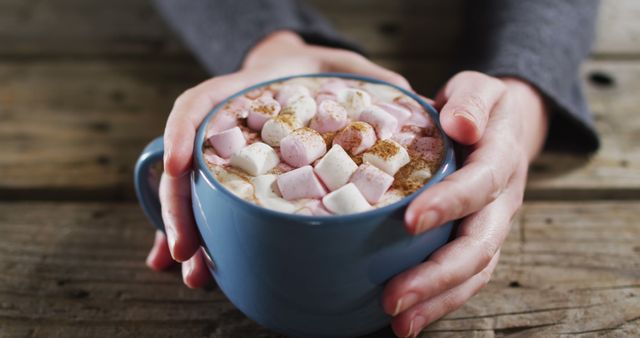 Close-up of hands holding blue mug filled with hot chocolate topped with marshmallows and sprinkled with cinnamon, on a rustic wooden table. Perfect for promoting cozy, winter-themed products, warm beverages, stress relief, and comfort during the colder months.