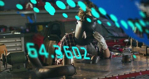 Woman welder wearing welding helmet and glove working at metalwork bench with pipes