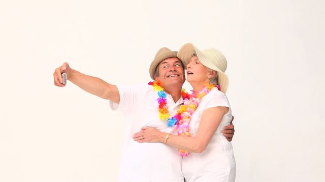 Cheerful mature couple wearing leis and summer hats while taking selfie. Shows joyful, carefree moment perfect for promoting vacation rentals, travel agencies, or photography products aimed at older audiences seeking excitement and rejuvenation.