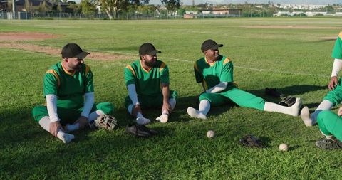Amateur baseball players sitting on grass stretching and laughing in green uniforms