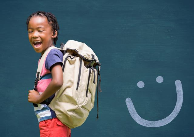Young African American boy wearing backpack, smiling in front of chalkboard with smiley face drawing. Ideal for back-to-school promotions, educational materials, and advertisements targeting elementary school students.