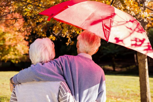 Senior couple embracing under a red umbrella in an autumn park, showcasing love and togetherness. Ideal for use in advertisements, articles, or websites focusing on elderly care, retirement, love in later life, and outdoor activities for seniors.