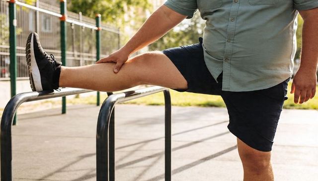 Overweight man stretching leg on parallel bars at sunny outdoor park for calisthenics