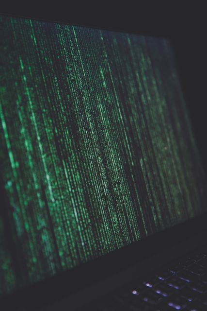 Digital concept image showing green binary code on a computer screen in a dark room. Useful for illustrating themes related to cybersecurity, hacking, programming, IT security, coding, and digital technology and encryption.